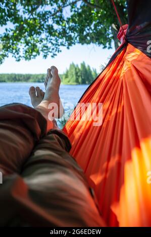 L'uomo a piedi nudi si rilassa disteso in un'amaca all'ombra degli alberi, con una magnifica vista sul lago, in una giornata estiva. Foto Stock