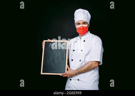 Cuoco cuoco che indossa giacca da cucina e cappello con maschera rossa, tenendo una lavagna con menu, su sfondo nero Foto Stock