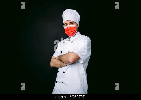 Cuoco cuoco che indossa giacca e cappello da cucina, con una maschera rossa, braccia incrociate, su sfondo nero Foto Stock