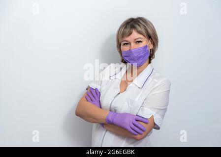 Infermiere in una uniforme medica in piedi con le braccia incrociate guardando la macchina fotografica e indossando camice bianco, maschera protettiva e guanti. Isolato su bianco Foto Stock