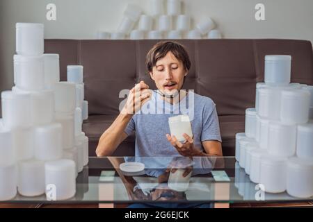 L'uomo mangia yogurt ed è circondato da molti vasi di yogurt che usa in un anno. Una grande quantità di vasetti di plastica per alimenti. Consumo eccessivo di Foto Stock