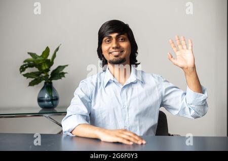 Bello vedere you.cheerful collega indiano agitando mano nella macchina fotografica, felice indù ragazzo in camicia formale si siede alla scrivania e saluto in linea interlocutore, prende una parte in conferenza online, webcam vista Foto Stock