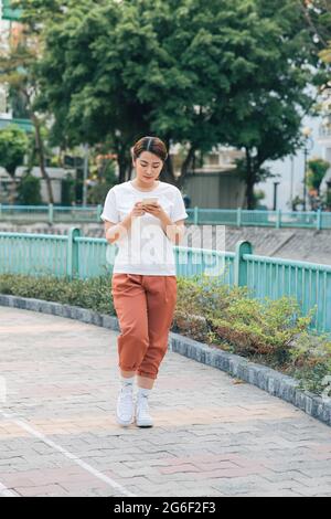 Giovane donna in abiti casual che cammina in un parco che chiacchiera sul suo telefono cellulare mentre sorride e aspetta. Foto Stock