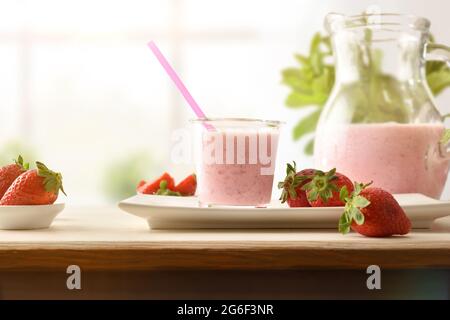 Bicchiere e brocca pieni di frullato di fragola con latte su panca di legno con finestra luminosa sullo sfondo. Vista frontale. Foto Stock