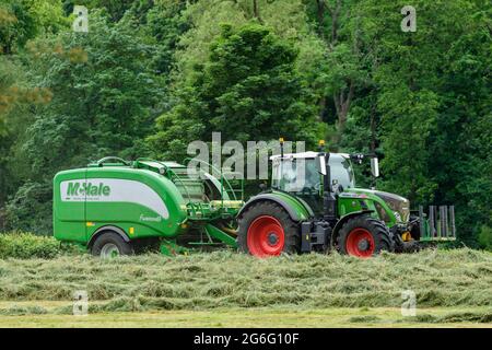 Produzione di fieno o insilato (agricoltore che guida un trattore agricolo verde sul campo, che tira l'imballatrice McHale, raccoglie e imballa l'erba secca) - Yorkshire Inghilterra UK. Foto Stock