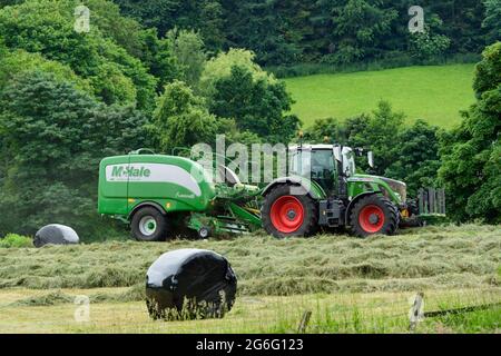 Produzione di fieno o insilato (coltivatore in trattore agricolo al lavoro in rotopressa di trazione di campo rurale, raccolta di erba secca e balle rotonde avvolte) - Yorkshire Inghilterra UK. Foto Stock