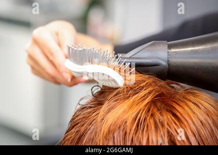 Closeup della mano del padrone con l'aria-asciugando e la spazzola che soffia i capelli rossi femminili in un salone Foto Stock