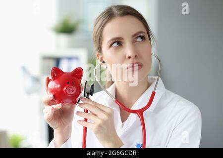 Il medico femminile in camice bianco tiene stetoscopio e lo applica alla banca di piggy Foto Stock