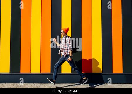 Foto di allegro positivo emozionante corsa saltando hipster uomo con valigetta cercando di successo contro isolato urbano multicolore muro sfondo Foto Stock