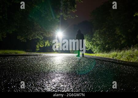 Un angolo basso, profondità di campo poco profonda di una figura con cappuccio e pozy, silhouette contro le luci della strada. Su una strada di campagna piovosa di notte. Foto Stock