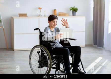 Un adolescente in sedia a rotelle con handicap positivo che ha una videoconferenza con l'insegnante su un tablet pc, agitando sullo schermo all'interno Foto Stock