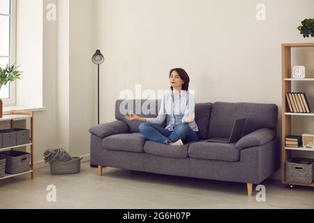 Donna d'affari che fa esercizi di yoga per alleviare lo stress mentre si siede sul divano in posizione lotus. Foto Stock