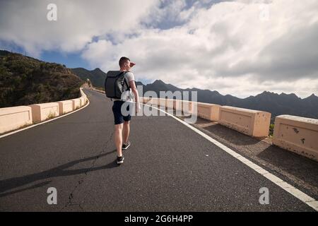 Vista posteriore dell'uomo con zaino su strada di montagna. Il turista giovane ama fare una gita in giornata di sole. Tenerife, Isole Canarie, Spagna. Foto Stock