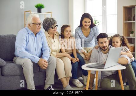 La famiglia si siede sul divano a casa e guarda i video sul computer portatile tutti insieme Foto Stock