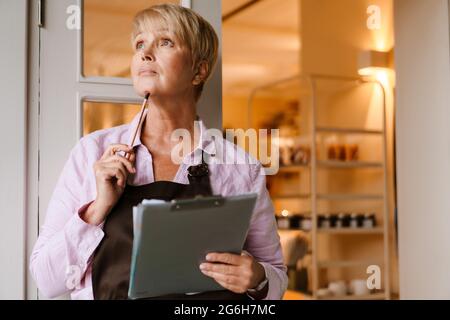 Donna matura che indossa grembiule in piedi con appunti nella porta del bar Foto Stock