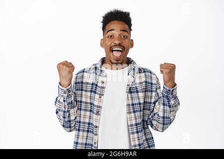 Felice africano americano giovane uomo festeggiando, vincendo, radunando per la squadra, clench e fare pugno pompa per vincere trionfo, raggiungere obiettivo, guardando stupito Foto Stock