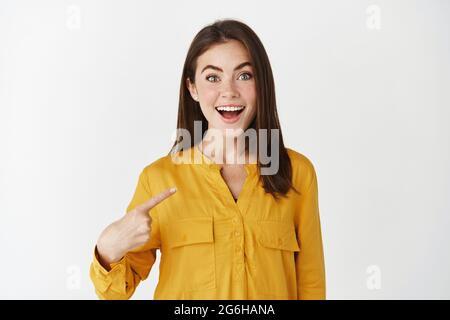 Donna felice e sorpresa che punta a se stessa, guardando stupita la macchina fotografica e sorridendo, essendo scelto, in piedi su sfondo bianco Foto Stock