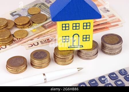Una pila crescente di monete in euro con una casa in cima, contanti, calcolatrice e penna. Una metafora finanziaria della Euro house. Foto Stock