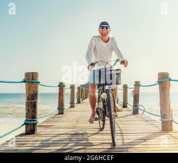 Ritratto di un uomo sorridente felice vestito con abiti estivi leggeri e occhiali da sole che cavalcano una bicicletta sul molo in legno. Vacanza senza cura in tropicale Foto Stock