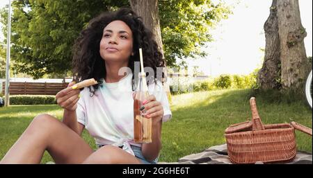 curly donna afroamericana che tiene fetta di pizza e bicchiere di limonata durante il picnic Foto Stock