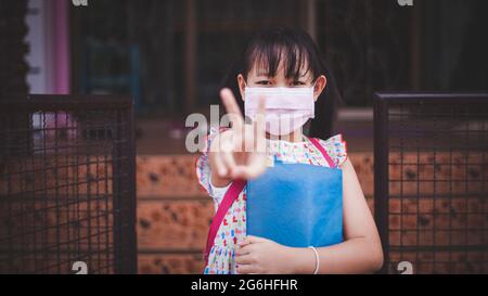 Ritratto di ragazza asiatica che indossa zaino e maschera facciale protettivo covid-19 pronto per la scuola elementare con la sua casa. Concetto di ritorno a scuola Foto Stock