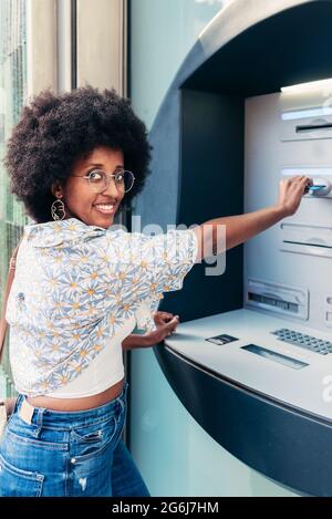 Ritratto verticale di una donna afroamericana che inserisce una carta di credito su un bancomat. Sta guardando la macchina fotografica un sorriso Foto Stock