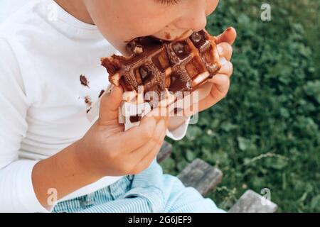 Ragazzo che mangia un waffle belga con un condimento al cioccolato. All'aperto. La vita quotidiana sporca macchia per lavare e pulire concetto. Foto di alta qualità Foto Stock