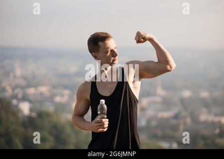 Bell'atleta maschile in abiti sportivi che mostra i suoi bicipiti muscolari mentre si posa in collina. Risultati di una formazione regolare e dura. Stili di vita attivi. Foto Stock