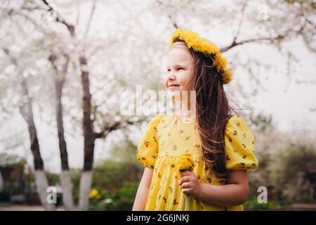 primo piano ritratto nel profilo di una ragazza sorridente in un vestito giallo e una corona di diandelioni gialli sulla testa con alberi senza foglie di primavera sul Foto Stock