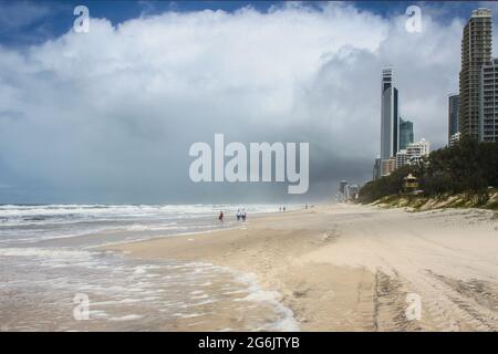 I turisti vagano sulla spiaggia di un mare tempesta con i grattacieli della Gold Coast dell'Australia che si aggirano in lontananza sotto un cielo nuvoloso sovrastato Foto Stock