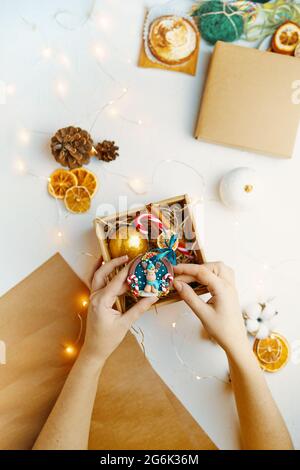 Vista dall'alto del souvenir natalizio in mano. Giocattolo realizzato in polimero creta.scatola regalo con decorazioni natalizie. Carta da imballaggio artigianale, coni, garland e arance secche su tavola bianca. Atmosfera di Capodanno. Foto Stock