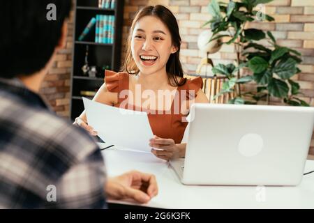 Giovane podcaster asiatico allegro che tiene un questionario mentre intervistando l'ospite maschile al posto di lavoro con microfono e computer portatile Foto Stock