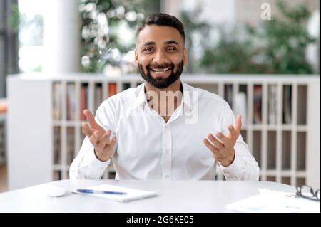 Amichevole positivo fiducioso indiano persona di affari o mentor, comunicante con il collega o cliente in chiamata di conferenza, guarda la macchina fotografica, gesturing con le mani, sorride piacevolmente, hanno intervista in linea Foto Stock