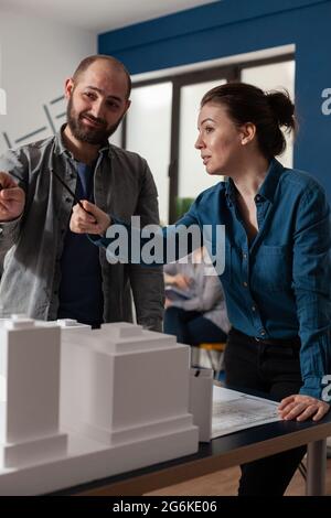 Il team di lavoro dell'architetto parla in un ufficio professionale in piedi alla scrivania con un modello di costruzione in maquette. Partner di progetto caucasici che cercano di progettare un progetto per il layout di costruzione Foto Stock
