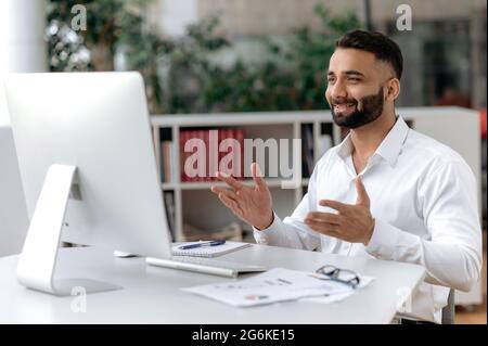 Un uomo d'affari indiano positivo di successo, ceo, siede a una scrivania in ufficio, comunica tramite videoconferenza con colleghi o partner, gesti con le mani, sorrisi, discute un nuovo progetto Foto Stock