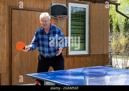 Senior maturo grigio caucasico vecchio uomo facendo palla forte colpo giocare ping pong tavolo tennis a casa giardino all'aperto in giornata di sole. Anziani Foto Stock