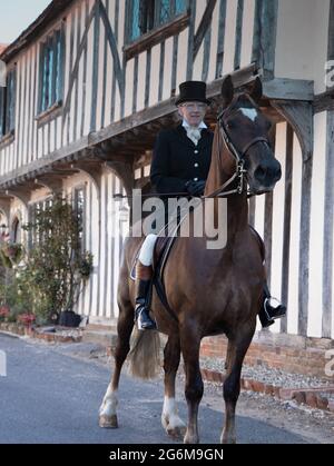 Lady formale di equitazione abitudine cavalcare un cavallo cacciatore di baia in un grazioso villaggio Suffolk di Nayland in Inghilterra con un vecchio edificio in legno sullo sfondo Foto Stock