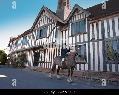 Lady formale di equitazione abitudine cavalcare un cavallo cacciatore di baia in un grazioso villaggio Suffolk di Nayland in Inghilterra con un vecchio edificio in legno sullo sfondo Foto Stock