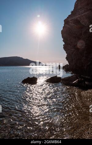 Sole che splende sulla spiaggia di ciottoli selvaggi sul Mar mediterraneo con silhouette di rocce scure, acqua limpida e fascio di sole. Viaggia in Grecia vicino ad Atene. Estate mare Foto Stock