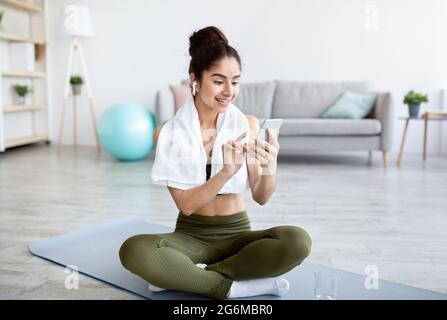 Affascinante giovane signora indiana con auricolari e telefono cellulare scelta playlist musicale per l'allenamento domestico Foto Stock