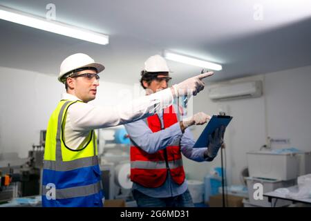 Ingegneri maschi adulti medi che esaminano la parte di macchina su una linea di produzione di maschera medica in una fabbrica. Foto Stock