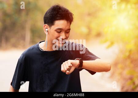 Un giovane sorridente sta facendo jogging nel parco. Sta guardando il suo orologio. Ascolta la musica sul suo telefono cellulare, la parte superiore del corpo si avvicina all'inginconia della fotocamera Foto Stock