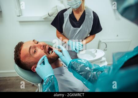 Maschio caucasico seduto nella stanza del dentista mentre l'infermiera aziona un dente rotto Foto Stock