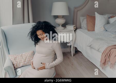 Amabile afroamericano godendo di buona gravidanza mentre si siede in camera da letto Foto Stock