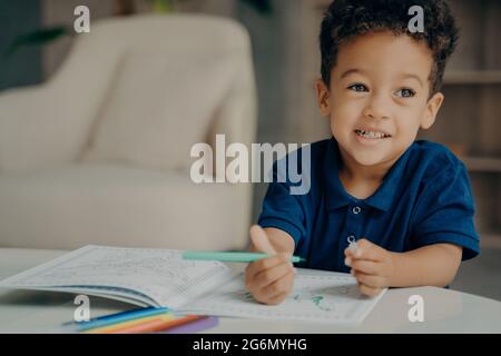 Carino afro americano piccolo ragazzo in blu scuro polo tshirt pittura libro da colorare a casa Foto Stock