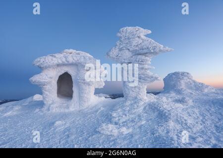 La casa è coperta di neve e ghiaccio di una textural misteriosa forma su un prato nevoso su uno sfondo di cielo blu. Fantastico paesaggio invernale. Foto Stock