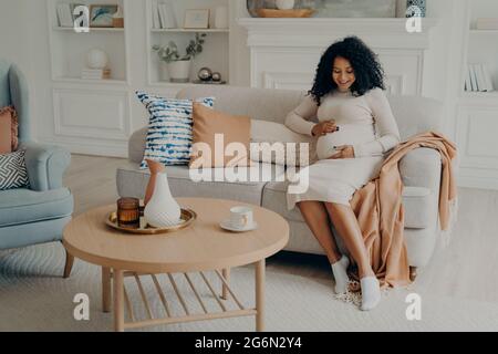 Donna afro americana incinta seduto sul divano e passare il tempo a casa Foto Stock