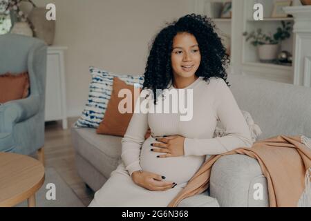 Sorridente bella giovane africana futura mamma seduta sul divano Foto Stock