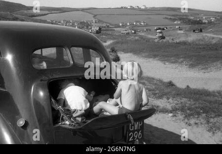 Anni '50, storico, due ragazzini nel bagagliaio o bagagliaio di un'auto dell'epoca, parcheggiato su alcune dune di sabbia, uno dei ragazzi nel deposito, l'altro esterno seduto sul portellone o sportello del bagagliaio ripiegabile, Inghilterra, Regno Unito. Foto Stock