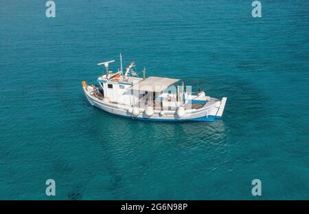Barca da pesca su sfondo mare turchese di colore blu. Vista aerea del drone. Peschereccio da traino tradizionale bianco e blu ormeggiato in acque ondulate dell'Egeo, giorno di sole. Foto Stock